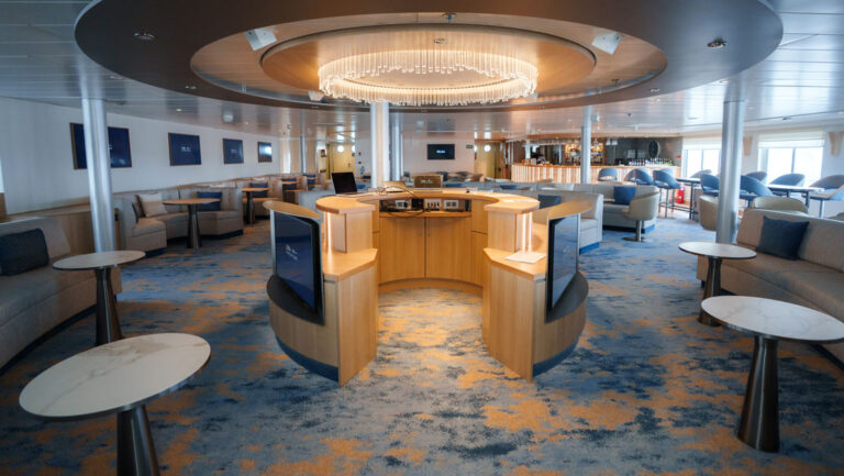 Interior lounge on National Geographic Explorer ship with round dais under chandelier, surrounded by padded benches & small tables.