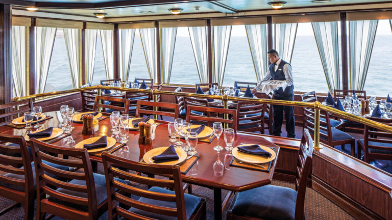 Crew member setting tables in dining room with window-lined walls aboard National Geographic Islander in Galapagos Islands