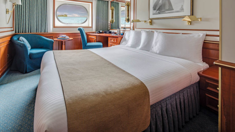 Category 2 cabin with large bed, armchair, small table, desk, chair and oval window aboard National Geographic Orion expedition ship