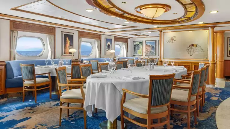 Dining room of Nat Geo Orion ship with tables set for dinner in white tablecloths, stemware, plates & blue booths along the wall.