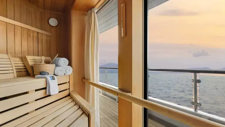 Sauna interior with beige wood bench & floor-to-ceiling windows looking onto sea at sunset aboard Nat Geo Orion.