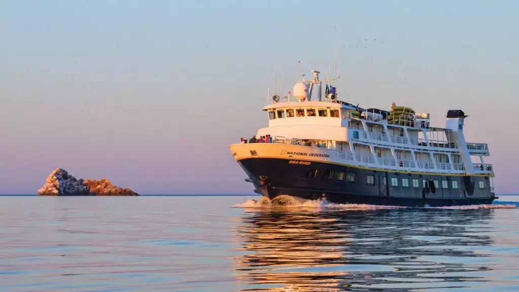 National Geographic Sea Bird small ship with dark blue hull & white upper decks cruises through calm seas at sunset.