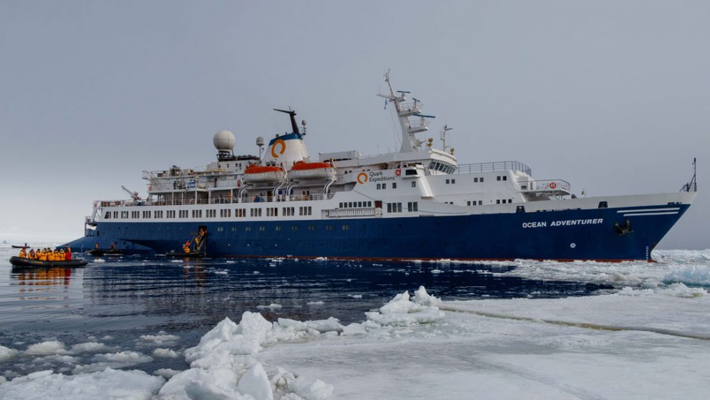 Starboard side of Ocean Adventurer ship with blue hull & white upper decks while parked by small icebergs.