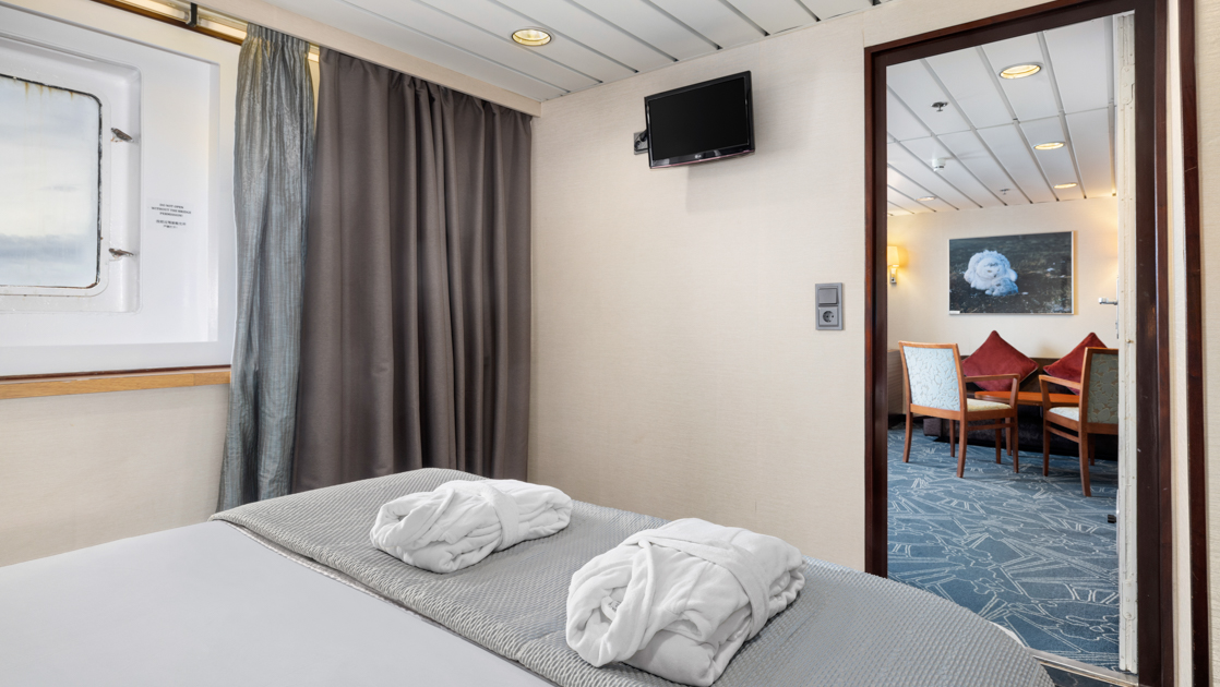 Junior Suite bedroom on Ocean Endeavour in Antarctica with double bed in white & silver linens with 2 folded bathrobes & window.