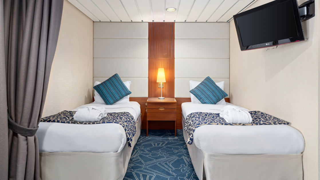 Category 3 cabin on Ocean Endeavour with 2 twin beds with teal throw pillows, tv & wood bedside table with lamp.