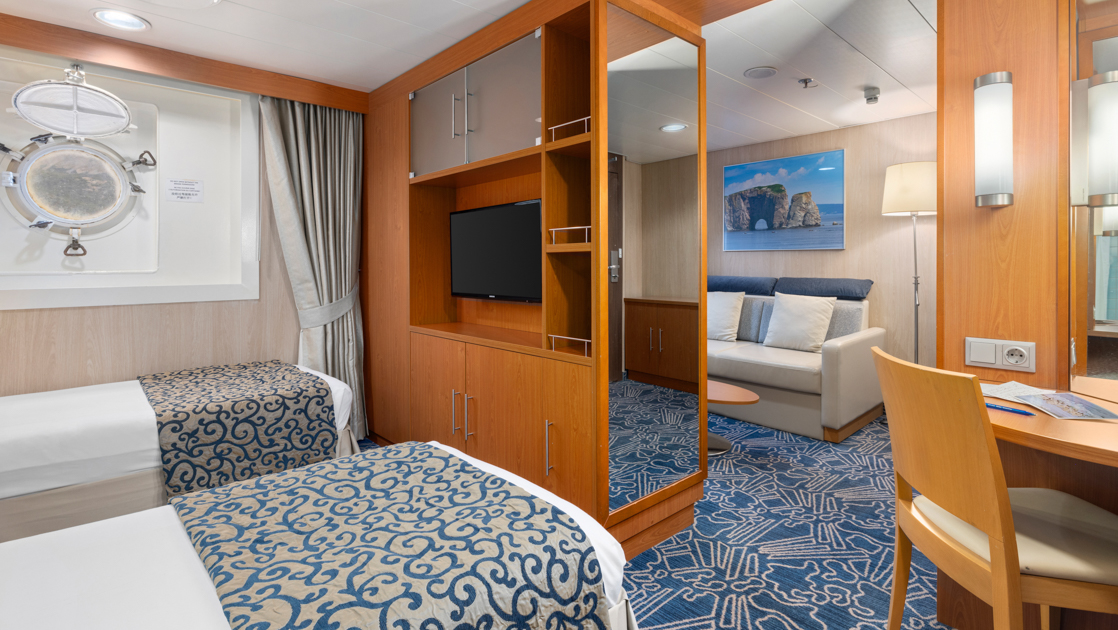 Category 6 cabin on Ocean Endeavour with porthole, wood desk & 2 twin beds partitioned from living room by wooden shelves & inset tv.