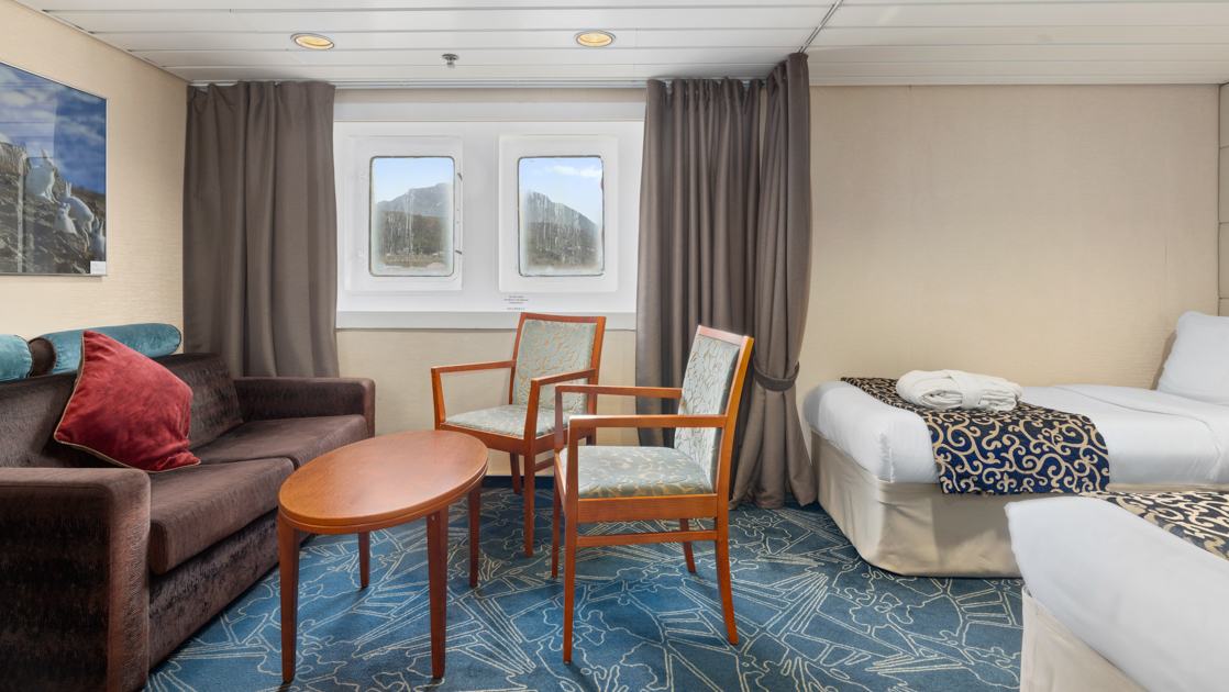 Category 8 cabin on Ocean Endeavour with 2 twin beds, 2 windows, brown upholstered couch & 2 wood chairs with blue cushions.
