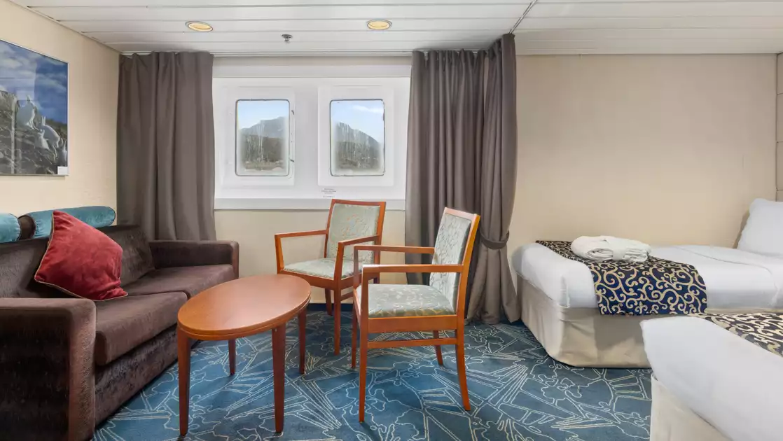 Category 8 cabin on Ocean Endeavour with 2 twin beds, 2 windows, brown upholstered couch & 2 wood chairs with blue cushions.