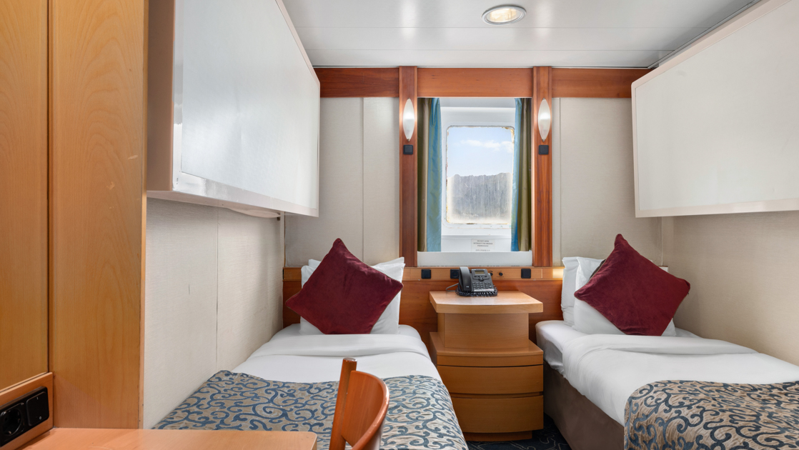Category 5 cabin on Ocean Endeavour vessel with 2 twin beds with red throw pillows, window, wood bedside table & desk.