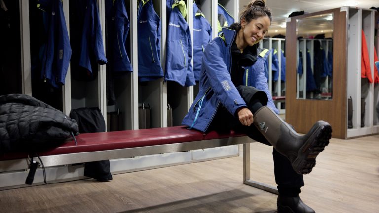 Woman in blue jacket sits on red bench & puts on muck boots in mudroom on Ocean Endeavour vessel, with open lockers behind.