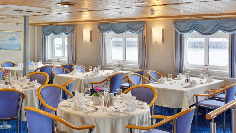 Dining room on Ocean Nova polar ship with view windows & 4- & 6-top tables set in white linens & upscale dinnerware.