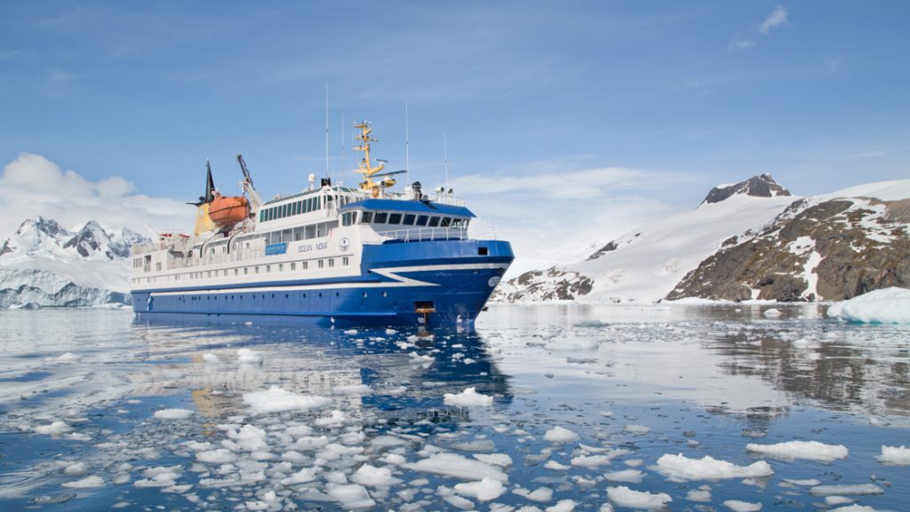 Ocean Nova polar ship with blue hull & white upper decks sits among iceberg bits on a sunny day in Antarctica.