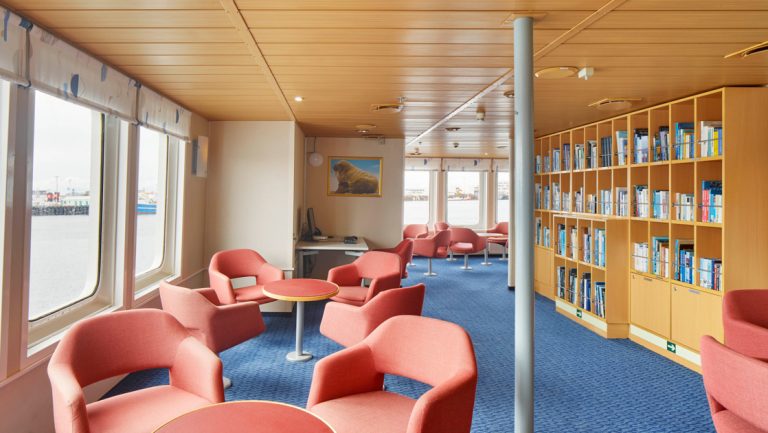 Library on Ocean Nova polar ship with red, round swivel chairs by fixed small tables & bookshelves beside view windows.