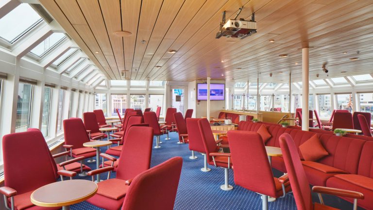 Observation lounge on Ocean Nova polar ship with red, round swivel chairs by fixed small tables & panoramic walls of windows.