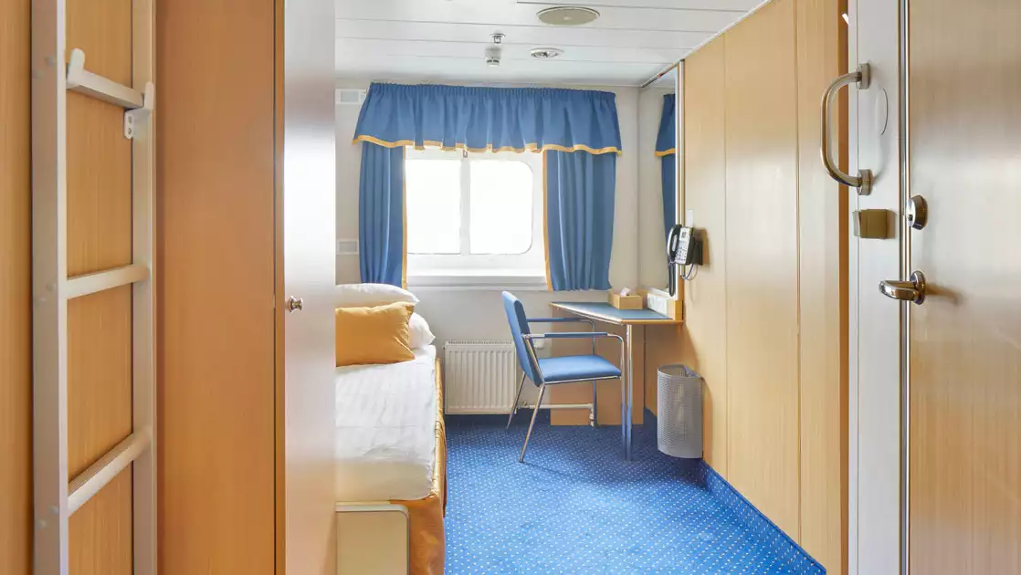 Twin bed in white linens in single cabin with blue & gold accents, small desk & wooden armoire on Ocean Nova ship.