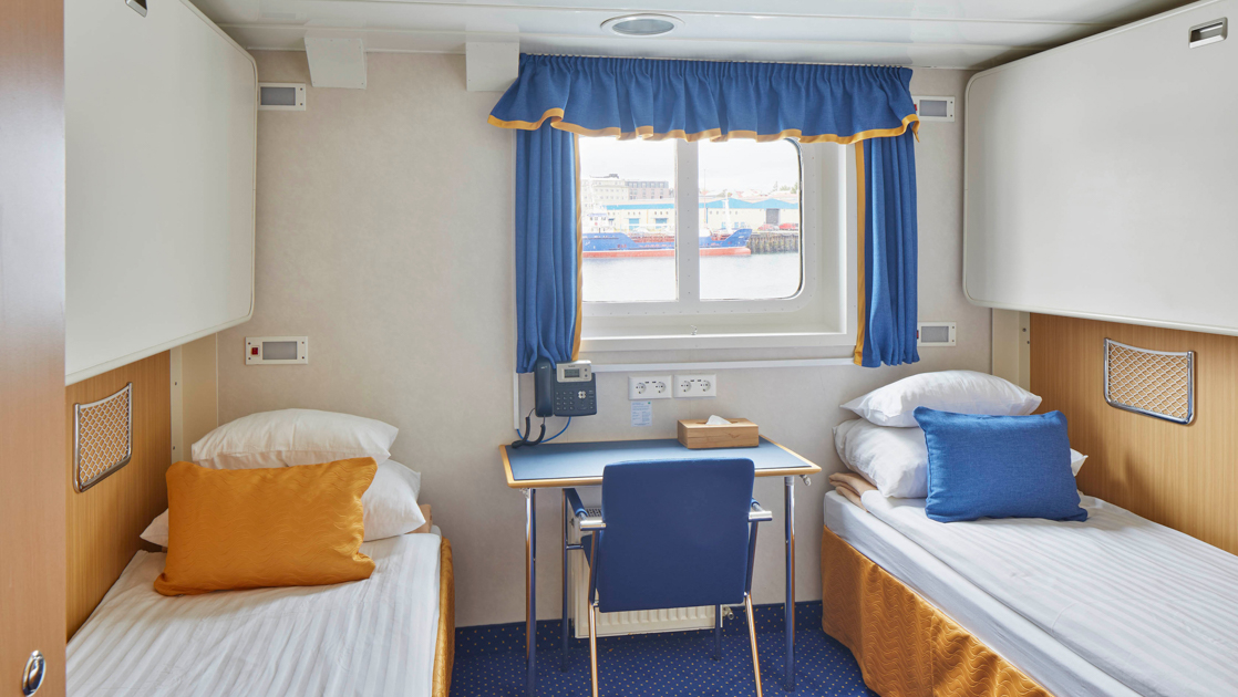 2 twin beds in white linens in twin cabin with blue & gold accents, small desk & wooden armoire on Ocean Nova ship.