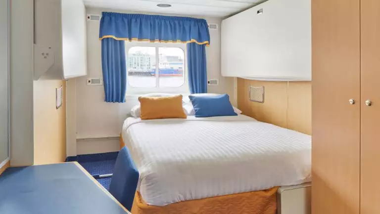 Double bed in white linens in twin cabin with blue & gold accents, small desk & wooden armoire on Ocean Nova ship.