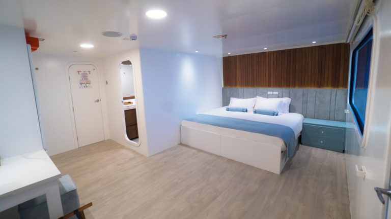 Large cabin with white walls, wooden accents & king-size bed covered in white & blue linens aboard catamaran Ocean Spray.