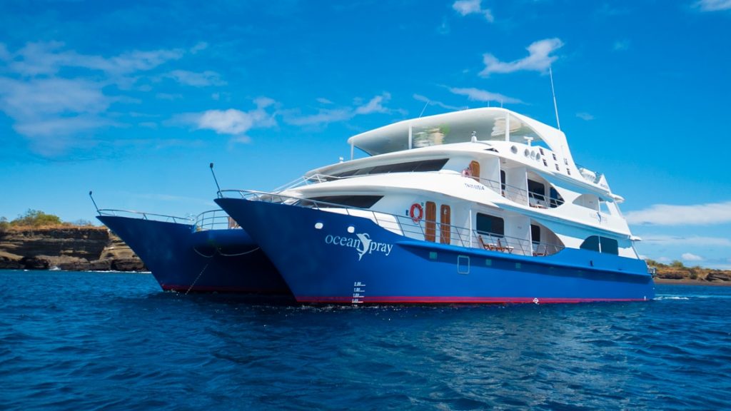 Small catamaran Ocean Spray with blue hull & white upper decks sits in calm water on a sunny day in Galapagos.