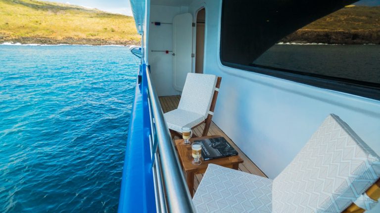 Narrow private balcony with 2 padded wooden chairs & wood coffee table on a ship sitting in calm water on a sunny day.
