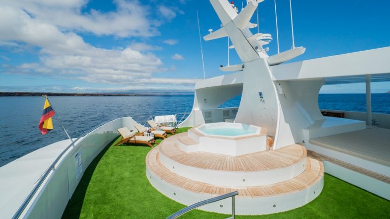 Small ship sun deck with bright green turf, raised Jacuzzi, Ecuadorian flag & chaise loungers under a bright blue sky.