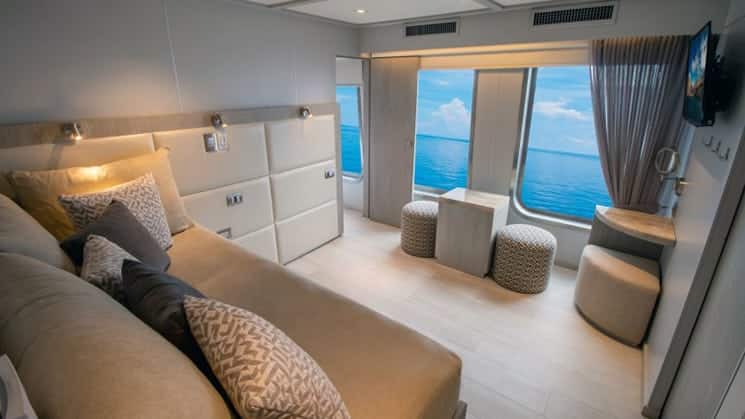 2-Bedroom Suite aboard Origin, Theory & Evolve luxury Galapagos yachts, with adjoining room showing beige couch with many pillows, floor-length windows, flatscreen TV & natural decor.