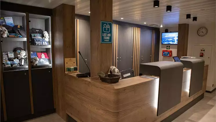 Modern reception area on Ortelius ship with light wood desk, white accent lights, beige floor & small giftshop in glass case.