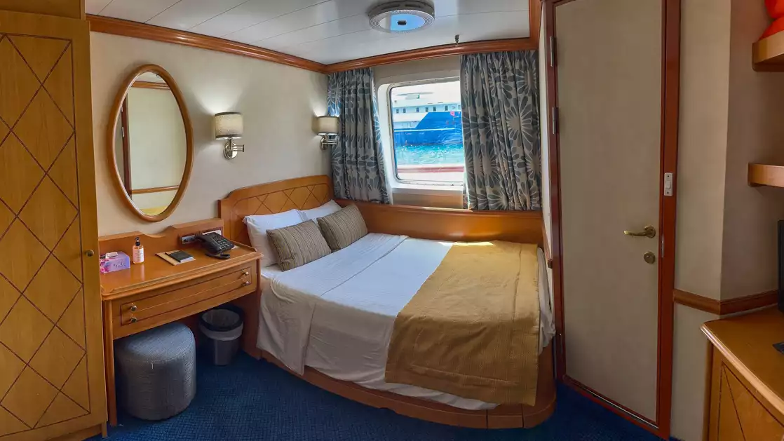 Double bed with mirror and wine and snacks on bed side table with small window aboard Panorama II