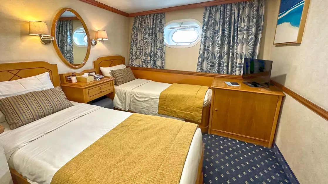 Two twin beds with towel made into a swan, mirror, porthole window and curtains aboard Panorama II.