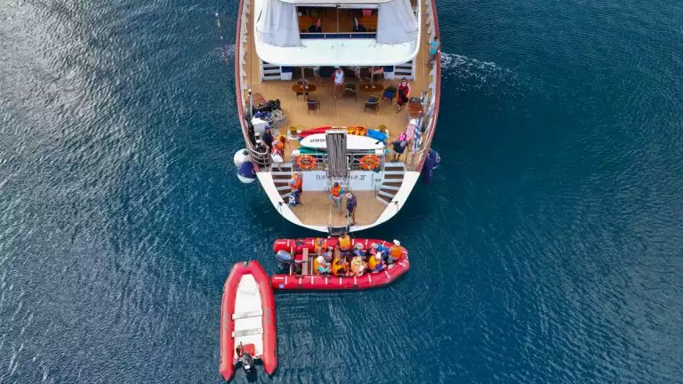 ariel view of the back of a sailboat with people on it and skiffs pulled up full of people ready to depart