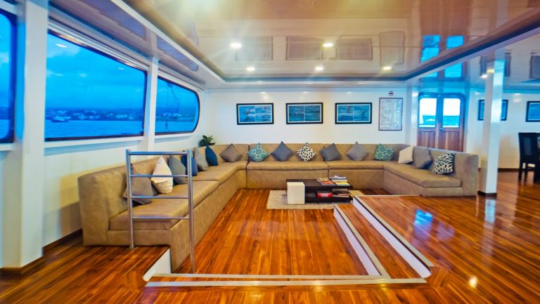 Living area on Petrel boat in Galapagos with sunken wood floor, wraparound beige couch with throw pillows & panoramic windows.