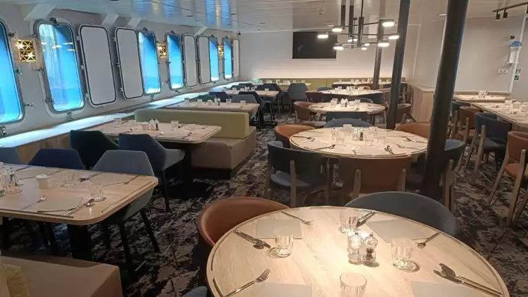 Circular & square light wood tables on expedition ship Plancius, set for a meal with dark blue & orange padded chairs & green padded booths.