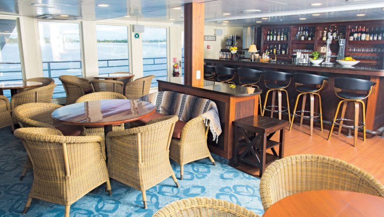 lounge on Safari Voyager small ship with wicker chairs, wood tables, granite bar, windows lining the room & blue carpet.