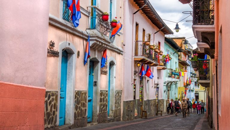 Colorful cobblestone street in Quito, Ecuador, with bright blue doors & multicolored flags flying from small window balconies.
