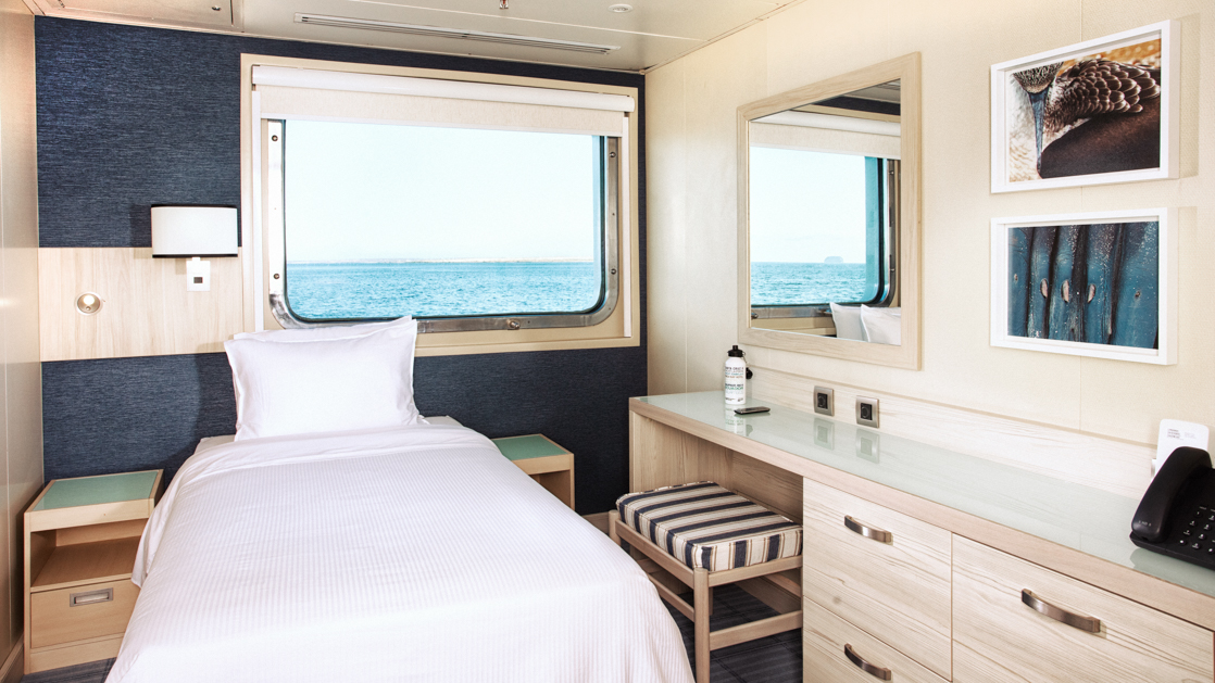 Twin bed with a large window above the bed featuring a vanity and dresser with blue accents aboard the Santa Cruz II Galapagos