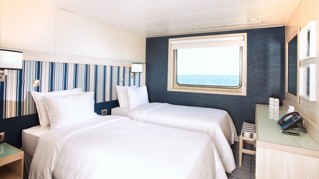 A beautiful room featuring twin beds a large window and dresser with blue accents aboard the Santa Cruz II in the Galapago Isles.
