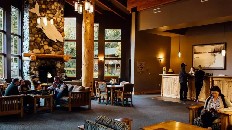 Large windows facing the mountain range. The main lodge at the Seward Windsong Lodge in Alaska couches and a stone fireplace.