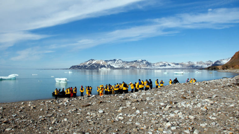travelers stand on the arctic shore where there is a zodiac inflatable skiff waiting to take them back to the small ship