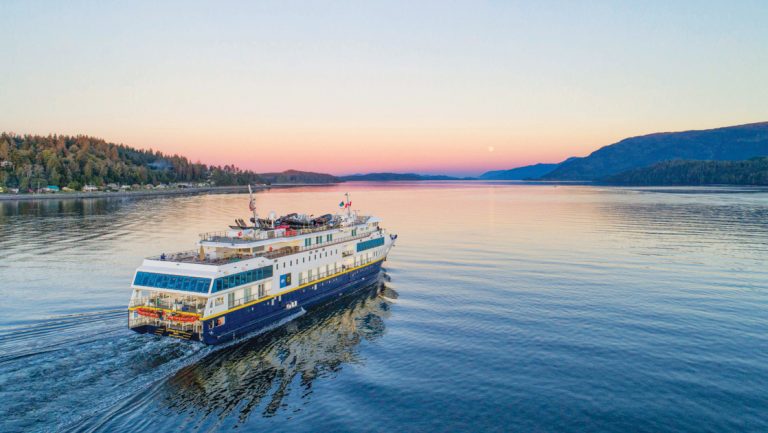 Small expedition ship with dark blue hull & white upper decks cruises in glassy water at sunset in the AK & BC inside passage.