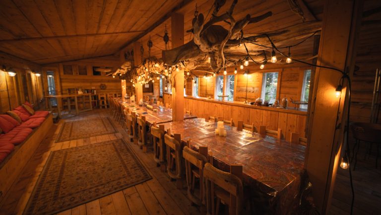 Widgeon boat house dining room at Tutka Bay Lodge with wood decor, long tables, antlers hanging, area rugs & red bench seating.