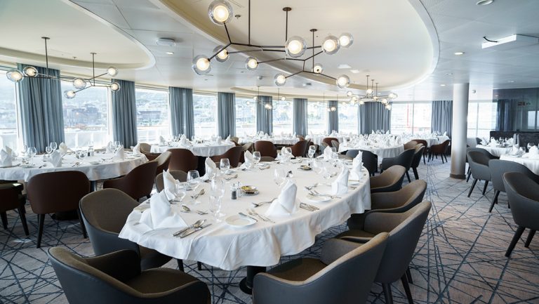 Dining room with modern decor, large tables in white tablecloths & fine dinnerware aboard Ultramarine ship.