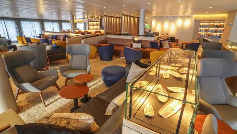 Lounge with modern swivel chairs, leather couches, view windows & glass-case artifacts on Ultramarine ship.