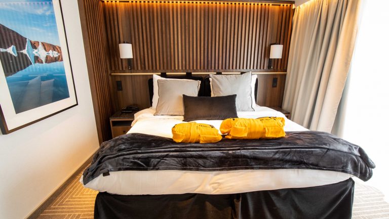 Double bed on Ultramarine ship with white sheets & duvet, gray fleece blanket, black bed skirts, wood headboard & large window.