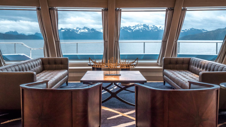 Two sofas and two chairs around a coffee table by windows in the lounge aboard Ventus Australis with views of the mountains out the windows.