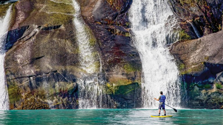 Man stands atop a paddleboard in turquoise water in front of tall waterfalls cascading over a large rock cliff wall in Alaska.
