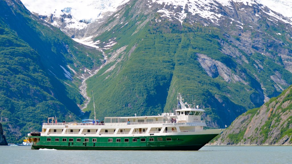 Wilderness Discoverer boat with forest green hull & 2 white upper decks cruises past green mountains with snow-covered peaks.
