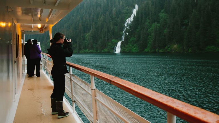 Woman in black pants & jacket looks through binoculars on side deck of Wilderness Explorer while cruising by a waterfall.