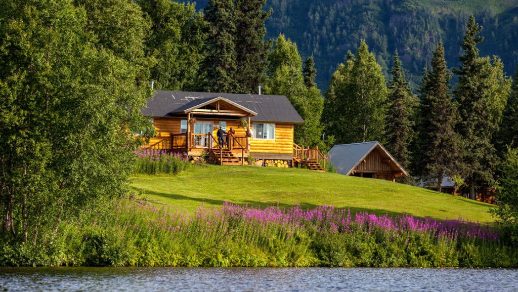 The Winterlake Lodge is perched on a grassy knoll above flowers and water, with pine trees in the background, in Alaska