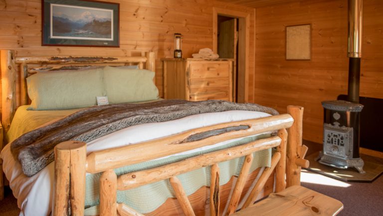 Wood post & beam double bed with sea green sheets & white comforter by small gas fireplace in a rustic wooden cabin.