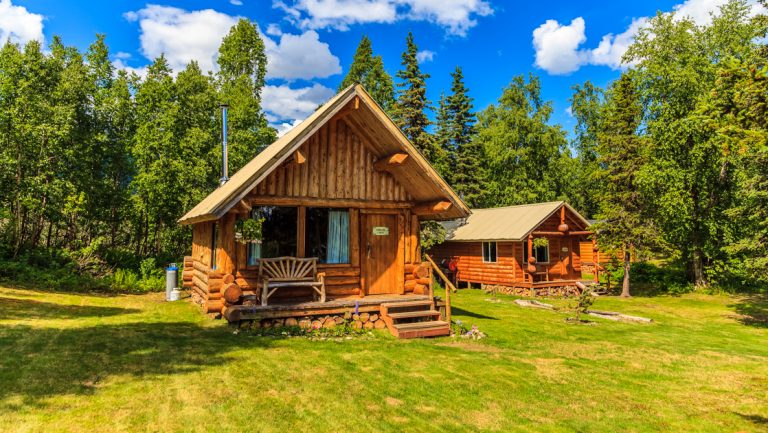Exterior of guest cabins at Winterlake Lodge with log cabin construction, front porch with a bench & grass & forest surrounding.